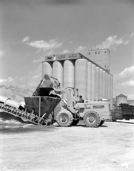 Man operating a Hough 90 Pay Loader to move sand or other material into conveying machinery. A sign on the driver's side door of a truck in the background reads: "Saginaw Asphalt Paving Co."