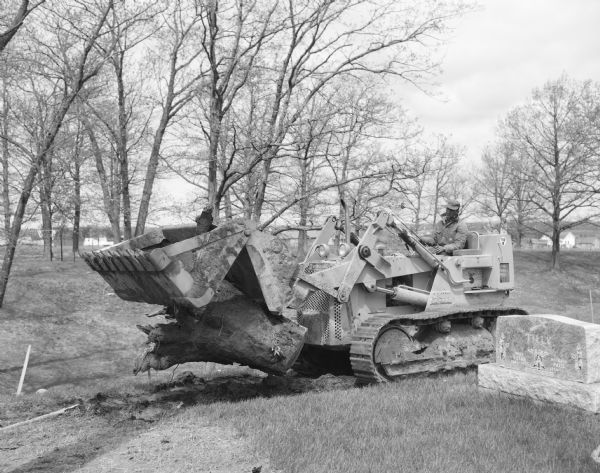 Man operating a Drott Skid-Shovel to move a tree trunk near a grave. Houses are in the background.
