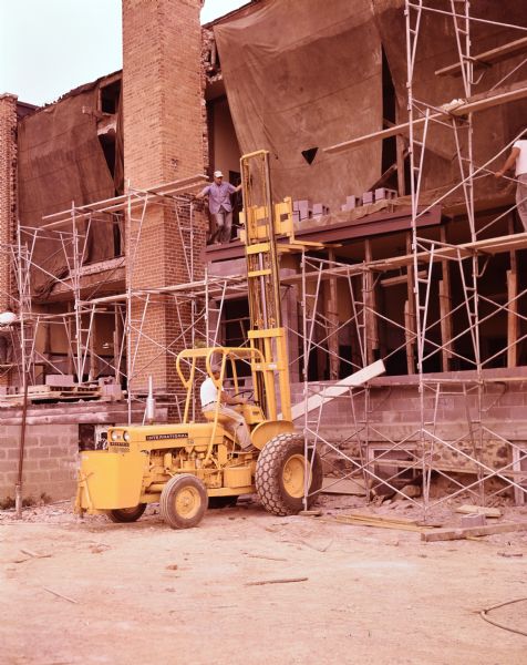 Man operating International construction equipment to raise building materials along the side of a brick building.