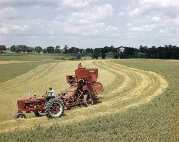 Elevated view of a man pulling a No. 122 Harvester Thresher with a Farmall M tractor. Trees and houses are visible in the far background.