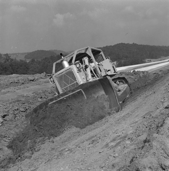 Three-quarter view from front left of a man operating a TD-25 on the side of a hill. There is a valley in the background. Vecellio and Grogan, Inc., working on highway project cutting four and one-half miles of Interstate through the rugged terrain of Brushy Mountain.