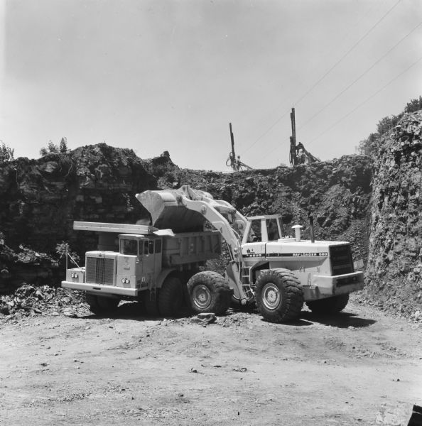 Man operating a Pay Loader to load a Pay Hauler. Other men are working on top of the hill being excavated. D.B. Hill contractors have contract to reconstruct 7.5 miles of route US-65. To move the rock, Hill is using International PH-140 Pay Haulers with a Hough 560 to do the loading.