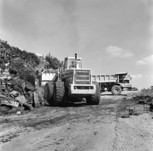 View up road towards a man operating a Pay Loader. A Pay Hauler is in the background. D.B. Hill contractors have contract to reconstruct 7.5 miles of route US-65. To move the rock, Hill is using International PH-140 Pay Haulers with a Hough 560 to do the loading.