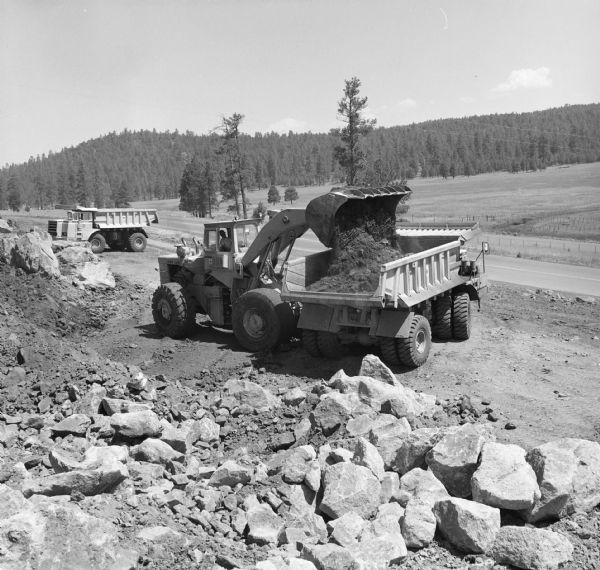 Elevated view of a man operating a Hough Pay Loader to load material into a 350 Pay Hauler. In the background is another Pay Hauler. Morrison-Knudsen Co., Inc. and Gordon H. Ball, Inc., a joint venture have a contract to construct south-bound lane of Interstate 17.