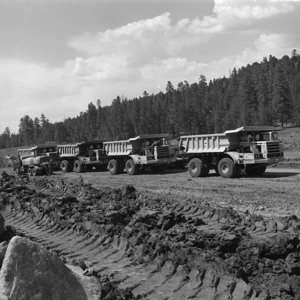 Four 350 Pay Haulers lined up in a row. There is a tanker truck parked on the left. Morrison-Knudsen Co., Inc. and Gordon H. Ball, Inc., a joint venture have a contract to construct south-bound lane of Interstate 17.