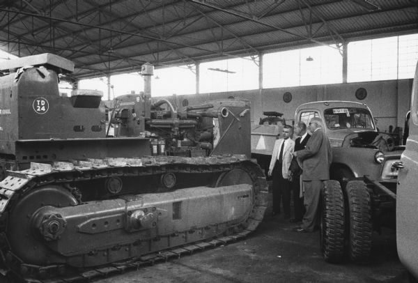 View of an International TD-24 and International trucks parked inside a building. International Harvester's resident representative in Colombia and Panama, Harry Perfect, is standing first from the right near two other men.