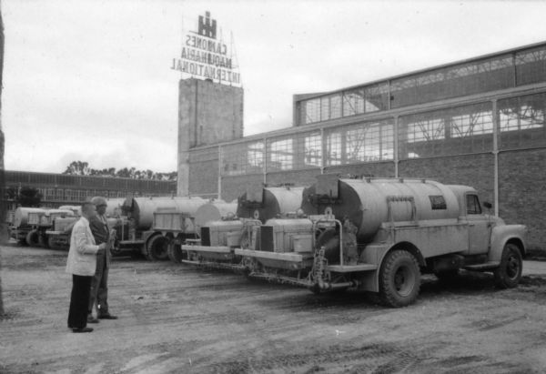 Two men standing outdoors near trucks parked at the International warehouse in Bogota. A large sign in the background, seen from the back, reads: "IH Camiones Maquinaria International."