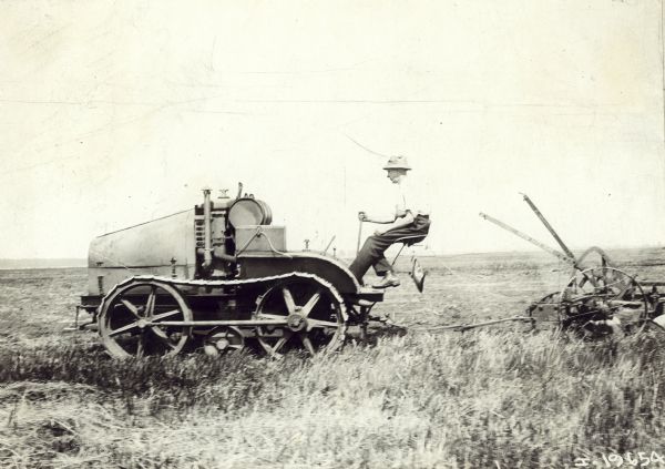 Left side view of a man driving an experimental tractor crawler in a field. There is an agricultural implement attached to the back of the tractor.