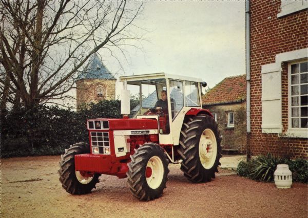 Color postcard view of a man posing in an International 1246 tractor in France. The tractor is parked in the yard next to a brick building.