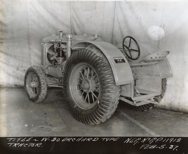 Three-quarter view from left rear of a W-30 Orchard type tractor. There is cloth canvas hung in the background as a backdrop. "Title — W-30 Orchard Type Tractor Neg. No. G.P. 11912 Feb. 5-37."