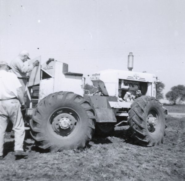 Two men are working outdoors in a field with The Big 4 tractor. One of the men is holding a panel that has been removed from the back of the tractor. Another man is standing on the ground in front of him.