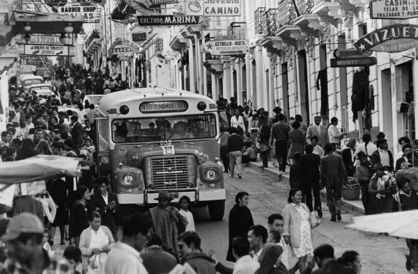 View up street towards the front of a Loadstar bus on a crowded street. Caption reads: "Crush of traffic on sign-studded street in Quito, Ecuador, includes IH bus. One of every three buses in Quito rides on IH chassis."