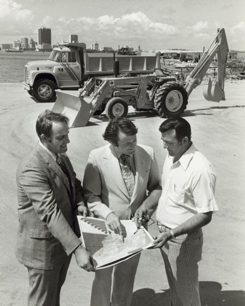 Slightly elevated view of three men standing outdoors and looking at a map. Behind them is a dump truck and a loader/backhoe. In the background is a city skyline. Accompanying public relations letter reads, in part: ... "International Harvester and its three distributors in Guatemala have teamed up to assist in the rebuilding of earthquake devastated sections of the country."