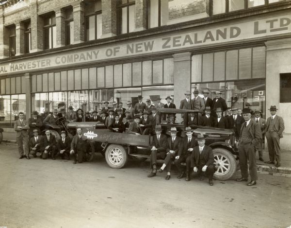 Large group portrait of men wearing suits and hats posing in front of the International Harvester Company's main office. There are large show windows on the first floor. The men are posing on and around a McCormick-Deering tractor and an International truck. The painted sign on the side of the truck reads: "A Size & Style for Every Business."