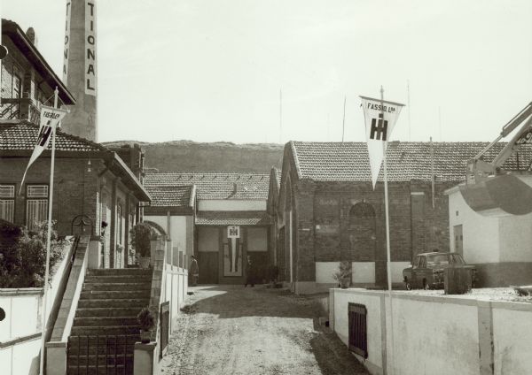 View of the buildings at Fassio, Lda.'s new premises in Portugal, Two men are walking near buildings near a banner with the IH logo. On the far right an automobile is parked near the side of a small building, and the raised shovel of a construction tractor is in the right foreground.