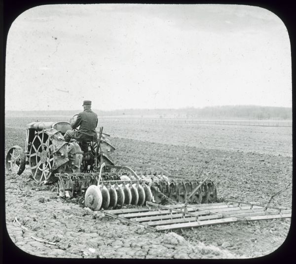 Three-quarter view from left rear of a man pulling a "combined disk and harrow" through a field with a tractor.