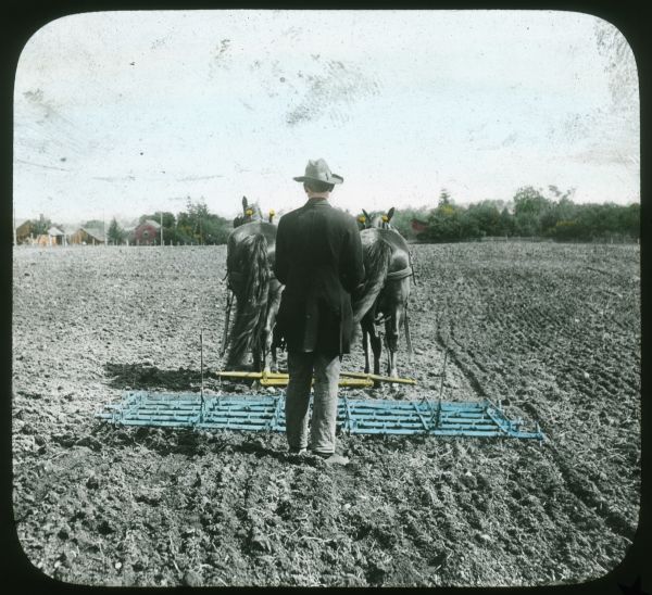 View towards a man walking behind a harrow in a farm field. Farm buildings are in the distance. Hand-tinted lantern slide.