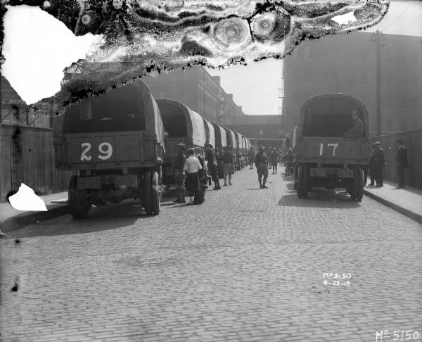 View down cobblestone street towards men, some on uniform, looking at trucks with army transport bodies parked on both sides of the street, near McCormick Works. The back of the truck on the left has the number "29" painted on it, and the one on the right is "17." Factory buildings are in the background, and tall wooden fences are along the sidewalks. These WWI era trucks are sometimes referred to as "Liberty Trucks."