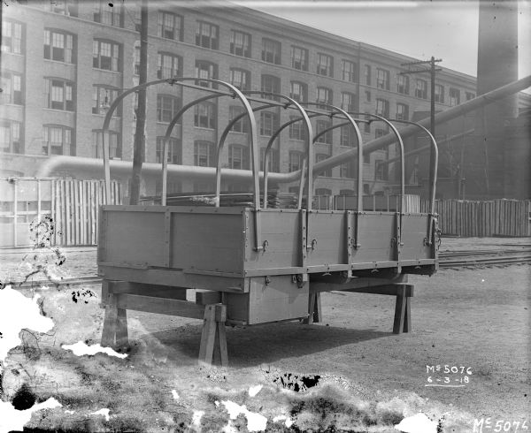 A truck frame for army use is set up on blocks in the yard at McCormick Works. The frame would be covered with a tarp. In the background are railroad tracks, a brick factory building, and a smokestack. These WWI era trucks are sometimes referred to as "Liberty Trucks."