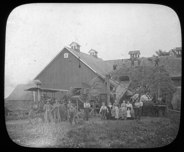 A large group of men, women and children are posing with threshing machinery, a steam engine and a horse-drawn wagon in front of a barn. Lantern slide. 