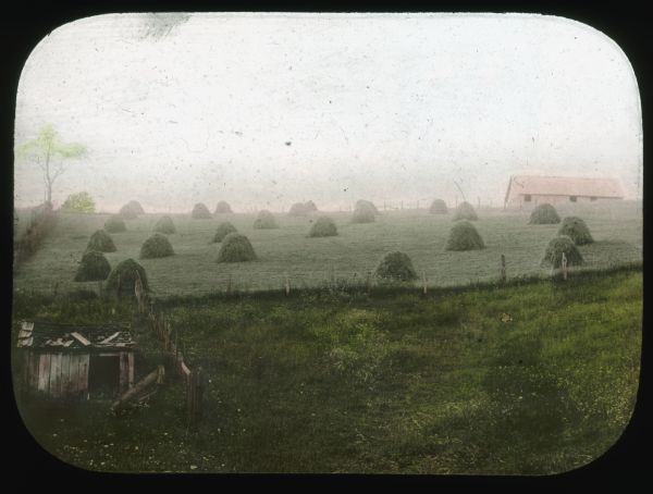 View towards a fenced field with stacks of freshly cut grass. A farm building is on the hill in the background on the right. Hand-tinted lantern slide.