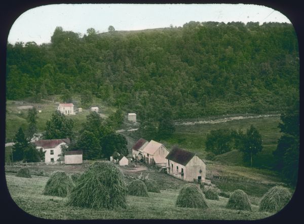 View down steep hill with haystacks. In the small valley below are farm buildings. Hand-tinted lantern slide.