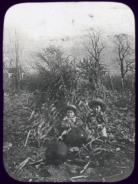 Two boys are sitting in a field, with pumpkins and corn on the ground in front of them, and a shock of corn behind them. Lantern slide. 