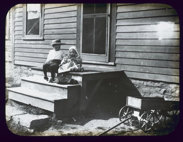 A boy and girl are sitting on the top wooden step of a house. There is a screen door behind them. A small wagon is in the right foreground. Lantern slide. 