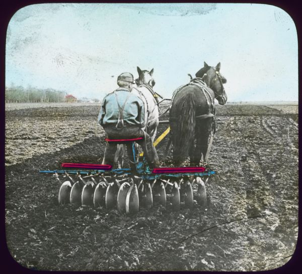 Rear view of a man sitting on a horse-drawn disc harrow working in a field. Hand-tinted lantern slide. 