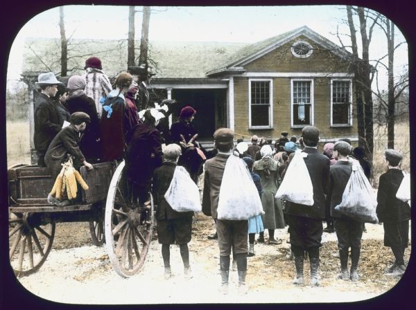 Rear view of a group of children, some holding sacks over their shoulders, and some standing on a horse-drawn cart, looking towards a building in the background. Hand-tinted lantern slide.