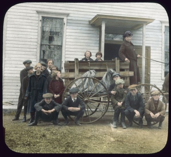 Boys and girls are standing and crouching next to a horse-drawn wagon. A woman is standing on the wagon holding the reins. There is a white, clapboard building behind the group. Hand-tinted lantern slide.