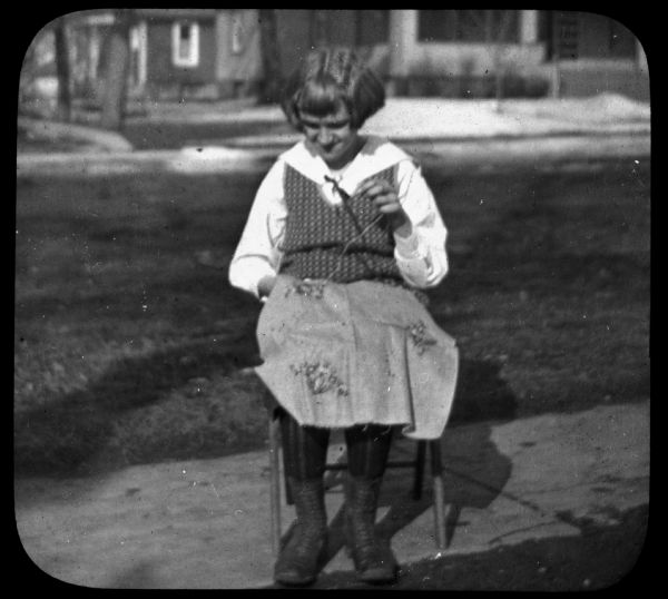 A girl is sitting on a chair on a sidewalk, sewing something resting in her lap. Buildings are in the background. Lantern slide.