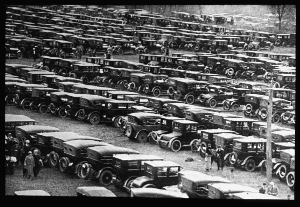 Elevated view of a large number of automobiles parked in rows in a field. People are walking and standing among the rows. Lantern slide.