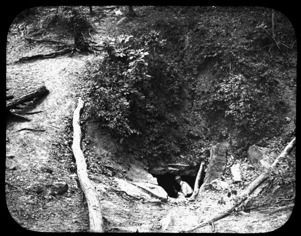 View looking down towards a man in the mouth of a cave on the side of a hill covered with plants and trees. Lantern slide.