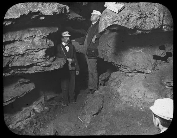 Two men are standing in a cave. They are both wearing hats that read: "El Dorado Natural Plastic." Another man is standing in the right foreground looking towards the two men. Lantern slide.