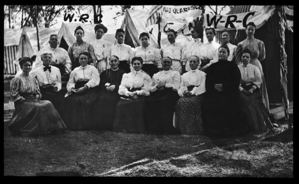 A group portrait of women sitting and standing in front of a row of tents. Some of the tents have signs that read: "Head Quarters" and "W.R.C." Lantern slide.