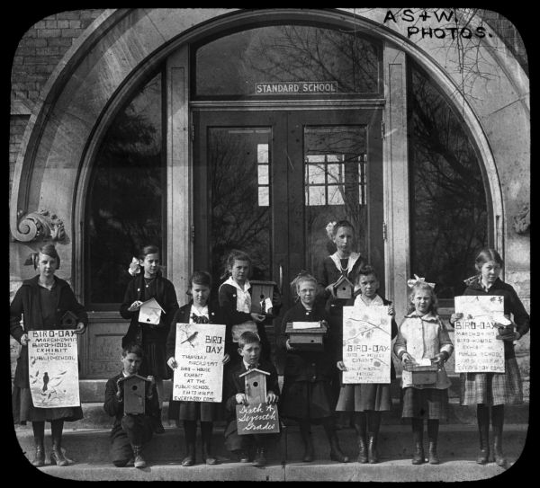 Outdoor group portrait of children standing in front of the Standard School. Each child is holding a birdhouse in their hands, and four of the children is also holding a hand-made "Bird-Day March 29, 1917" poster. One boy in the center is holding a sign that reads: "Sixth & Seventh Grades." Lantern slide.