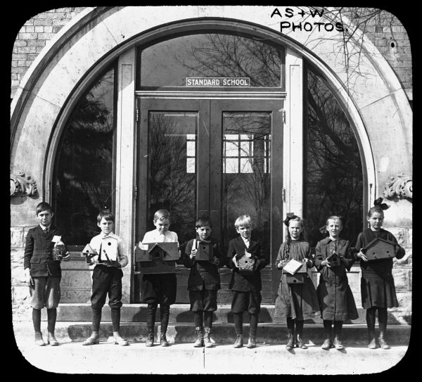 Outdoor group portrait of eight children standing in a row in front of the Standard School. Each child is holding a birdhouse in their hands. Lantern slide.