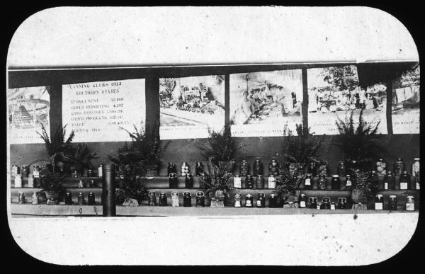 Canned vegetables are displayed on a table, with signs and illustrations hanging on the wall behind. One sign includes the text: "Canning Clubs 1913, Southern States." Lantern slide.