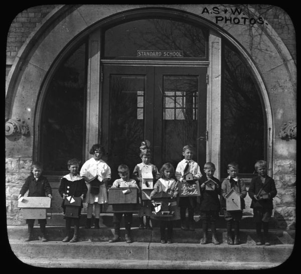 Outdoor group portrait of children standing in front of the Standard School. Each child is holding a birdhouse in their hands, and one of the children in the center is holding a sign that reads: "First Grade." Lantern slide.