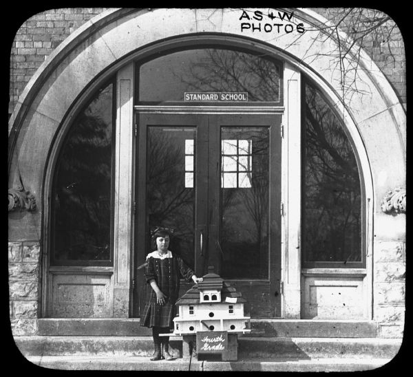 A young girl is standing with a large birdhouse in front of the large, arched doorway of the Standard School. The sign on the birdhouse reads: "Fourth Grade." Lantern slide.