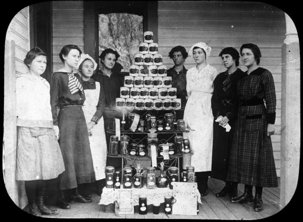 Group portrait of young women standing on a porch around a shelf displaying canned vegetables, and jars of vegetables. Some of the jars have ribbons on them. Lantern slide.