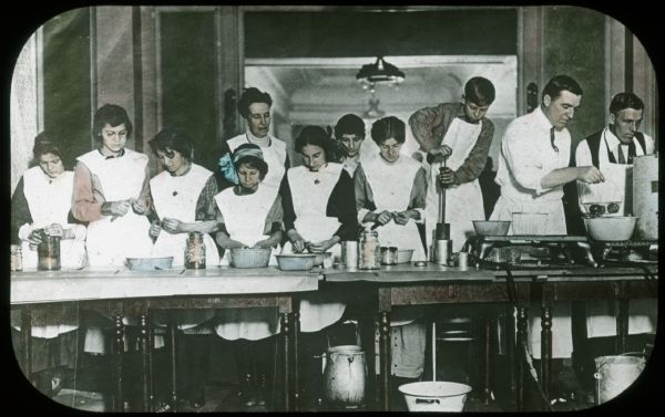 View towards a group of men, women and children working at tables with canning equipment. Lantern slide.