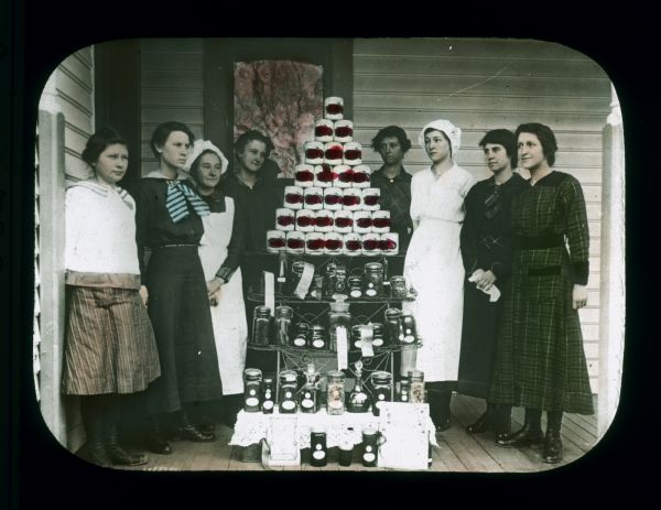 Group portrait of young women standing on a porch around a shelf displaying canned vegetables, and jars of vegetables. Some of the jars have ribbons on them. Hand-tinted lantern slide.