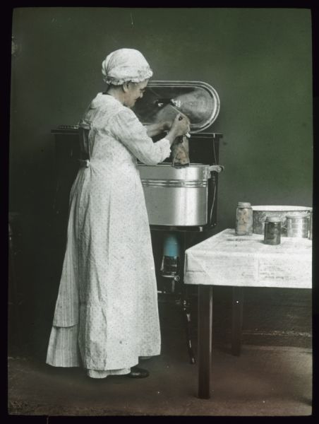 A woman wearing a white apron and hat working with home canning equipment. Hand-tinted lantern slide.