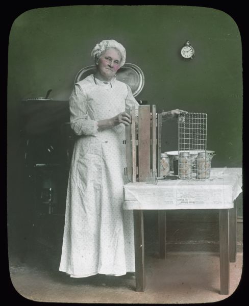 A woman wearing a white apron and hat is standing in front of a stove holding home canning equipment displayed on a table with canning jars.  Hand-tinted lantern slide.