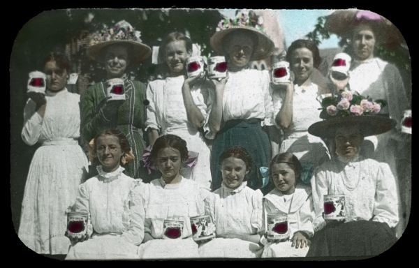 Group portrait of women and girls standing and sitting outdoors. They are each holding a can of IH Brand canned tomatoes. Hand-tinted lantern slide.