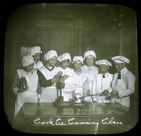 Group of young men and women in a canning class working indoors. Hand-tinted lantern slide.