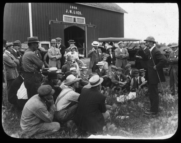 A man is standing and speaking to a group of men and women. They are gathered outdoors, and a sign on a barn above the door in the background reads: "1906 J.M. Lies." Lantern slide.
