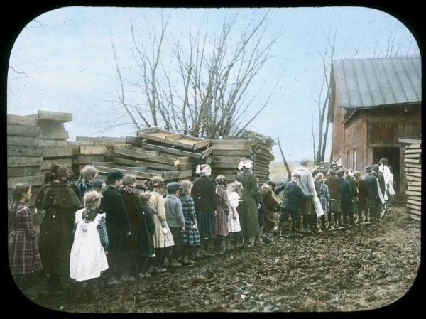A group of children are standing in line outdoors. They are on a wooden walkway leading to a farm building. There is mud in the driveway. Pallets are stacked on the left and right. Hand-tinted lantern slide.